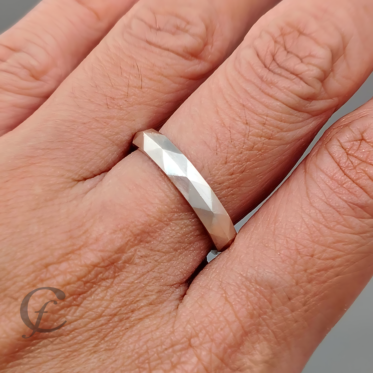 A close-up of a hand wearing a faceted silver band ring on the ring finger, with a soft gray background.