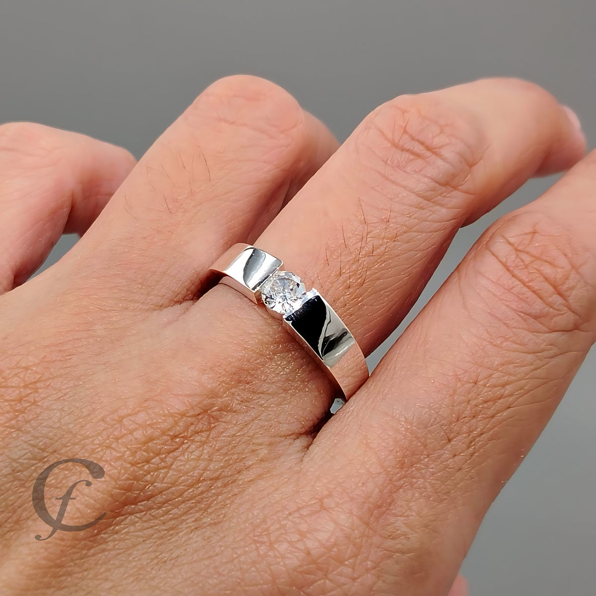 A polished silver band ring with a single round-cut clear gemstone, shown worn on a person's finger against a neutral gray background.