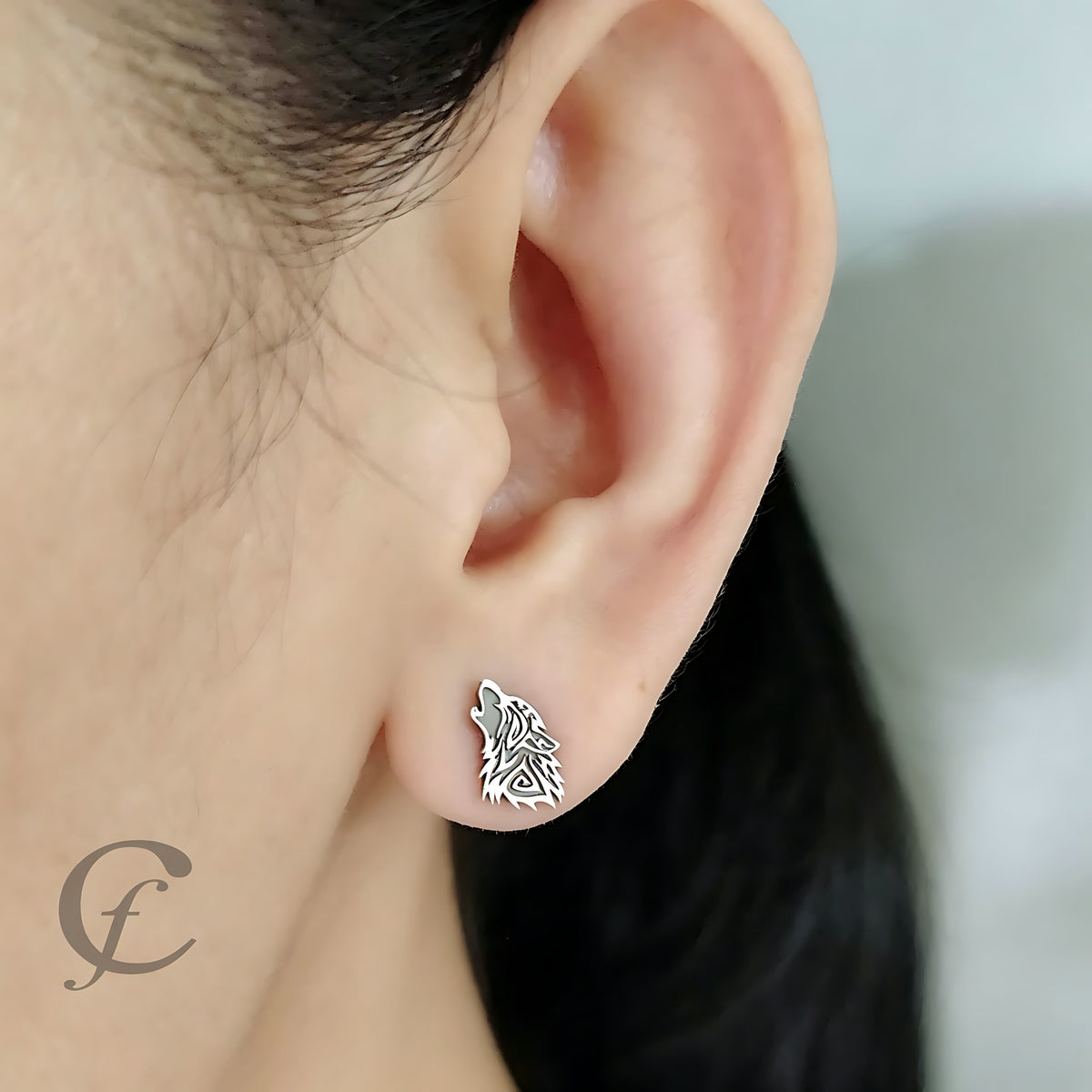 A close-up of a person's ear wearing a small silver wolf stud earring. The earring features an intricate, textured wolf head design with openwork details.
