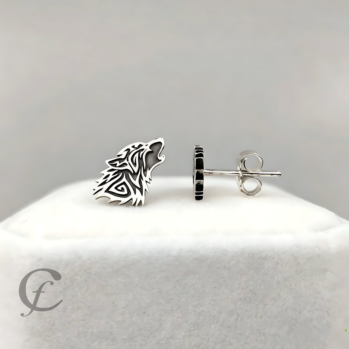 A pair of silver stud earrings displayed on a white jewelry stand against a light gray background. One earring is shown from the front, featuring a stylized wolf head cutout design, and the other is shown from the side, revealing a circular setting with small black inlay details and a silver butterfly back.