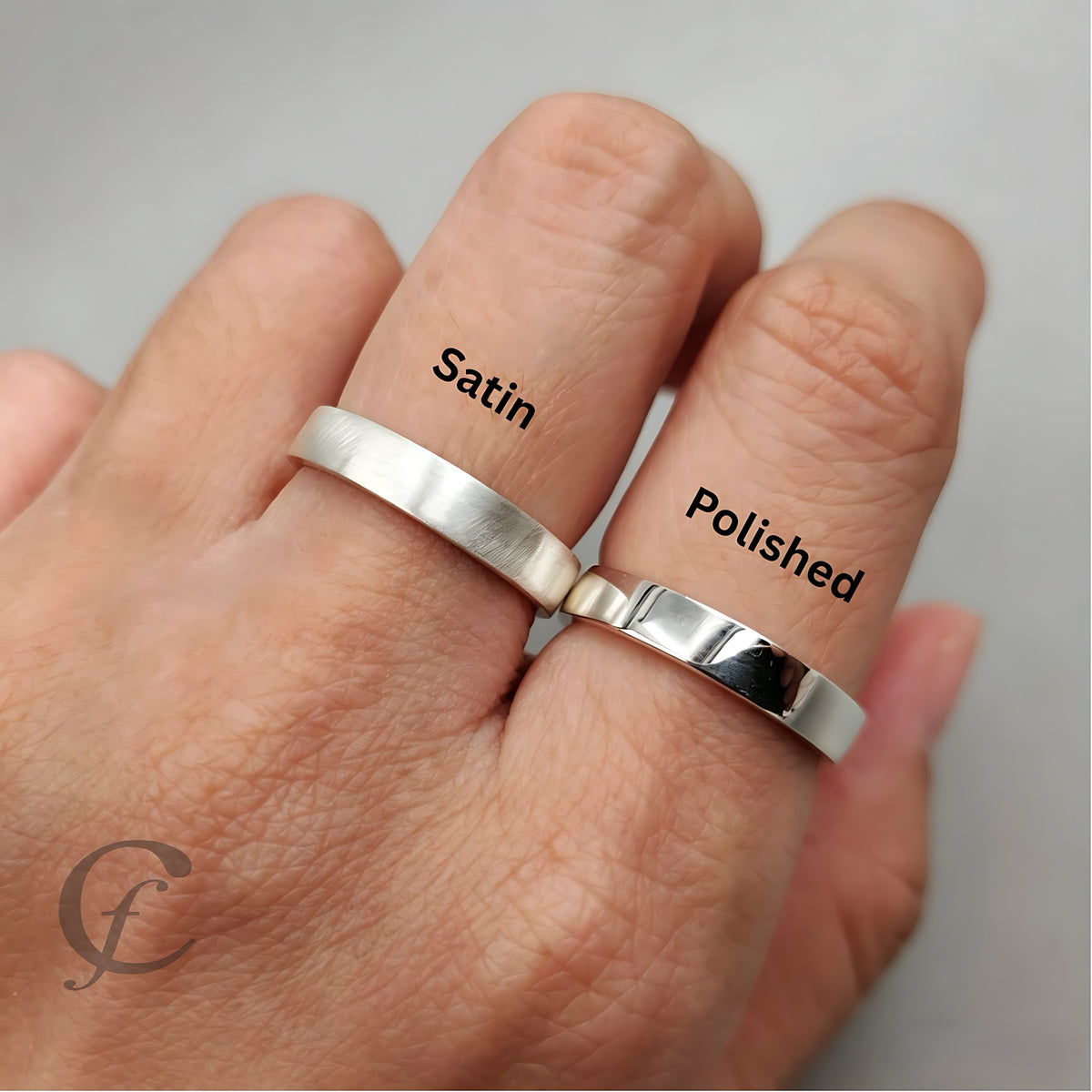 A close-up of a hand wearing two silver bands, labeled 'Satin' and 'Polished', to show the different surface finishes. The background is a soft, neutral gray.