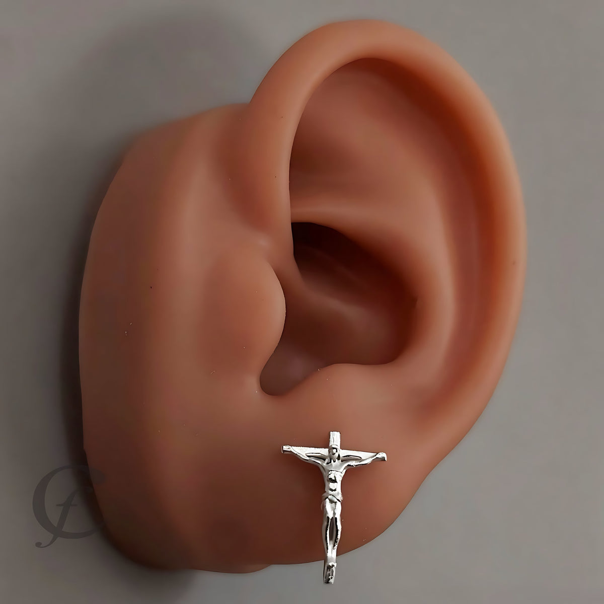 A silver crucifix stud earring shown on a realistic peach-colored ear model against a plain gray background.