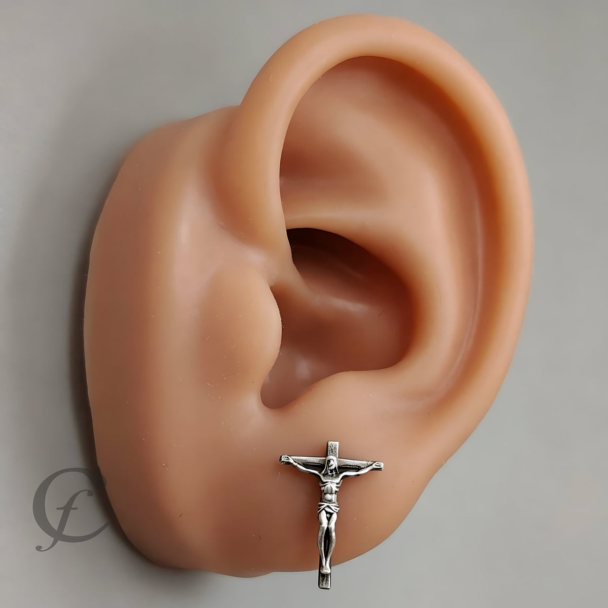 A silver crucifix stud earring displayed on the lobe of a realistic peach-colored ear model against a neutral gray background.