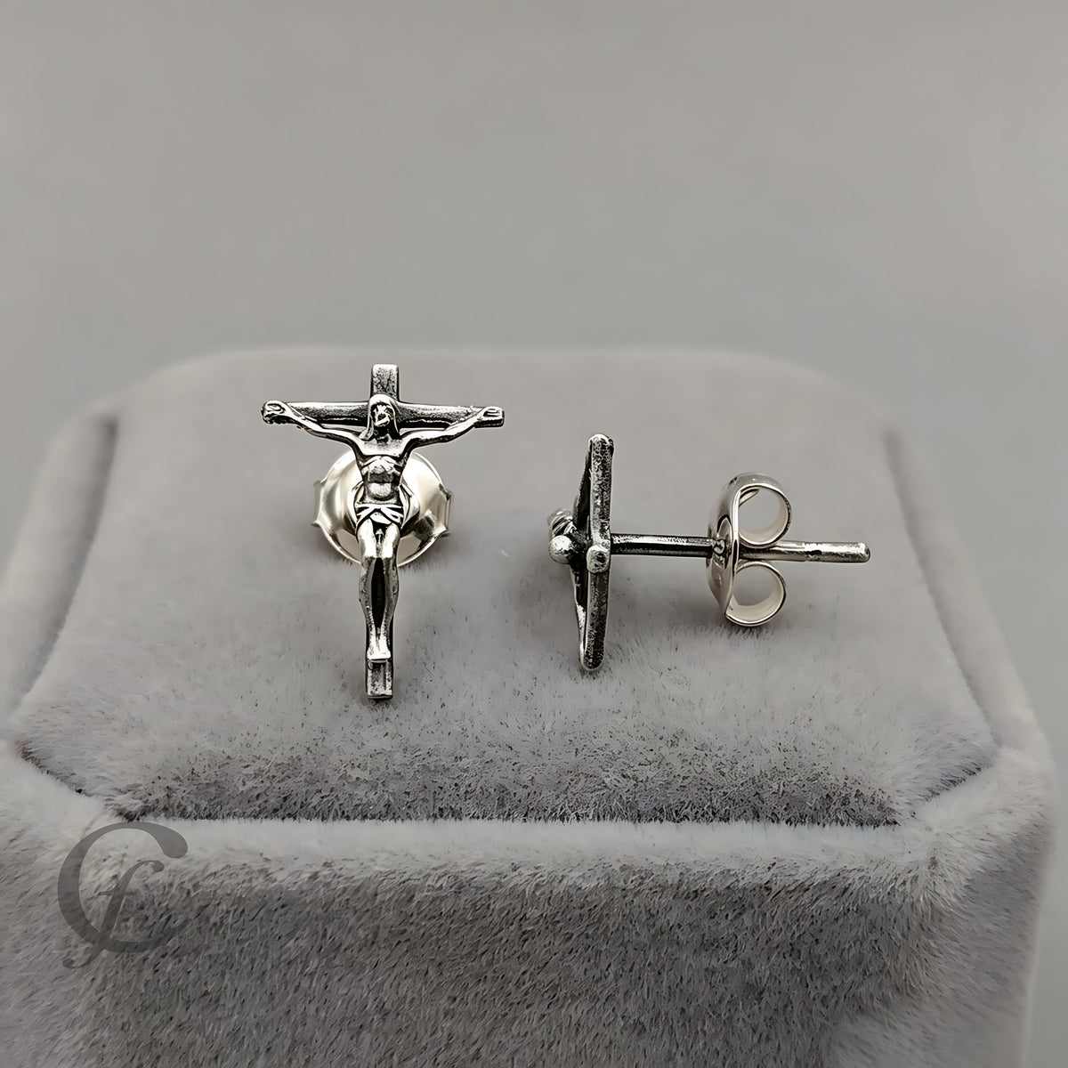 A pair of silver crucifix stud earrings displayed on a gray velvet jewelry box. One earring is shown from the front, featuring a detailed silver figure of Jesus on a cross, while the other is shown from the side/back, displaying the post and butterfly back.