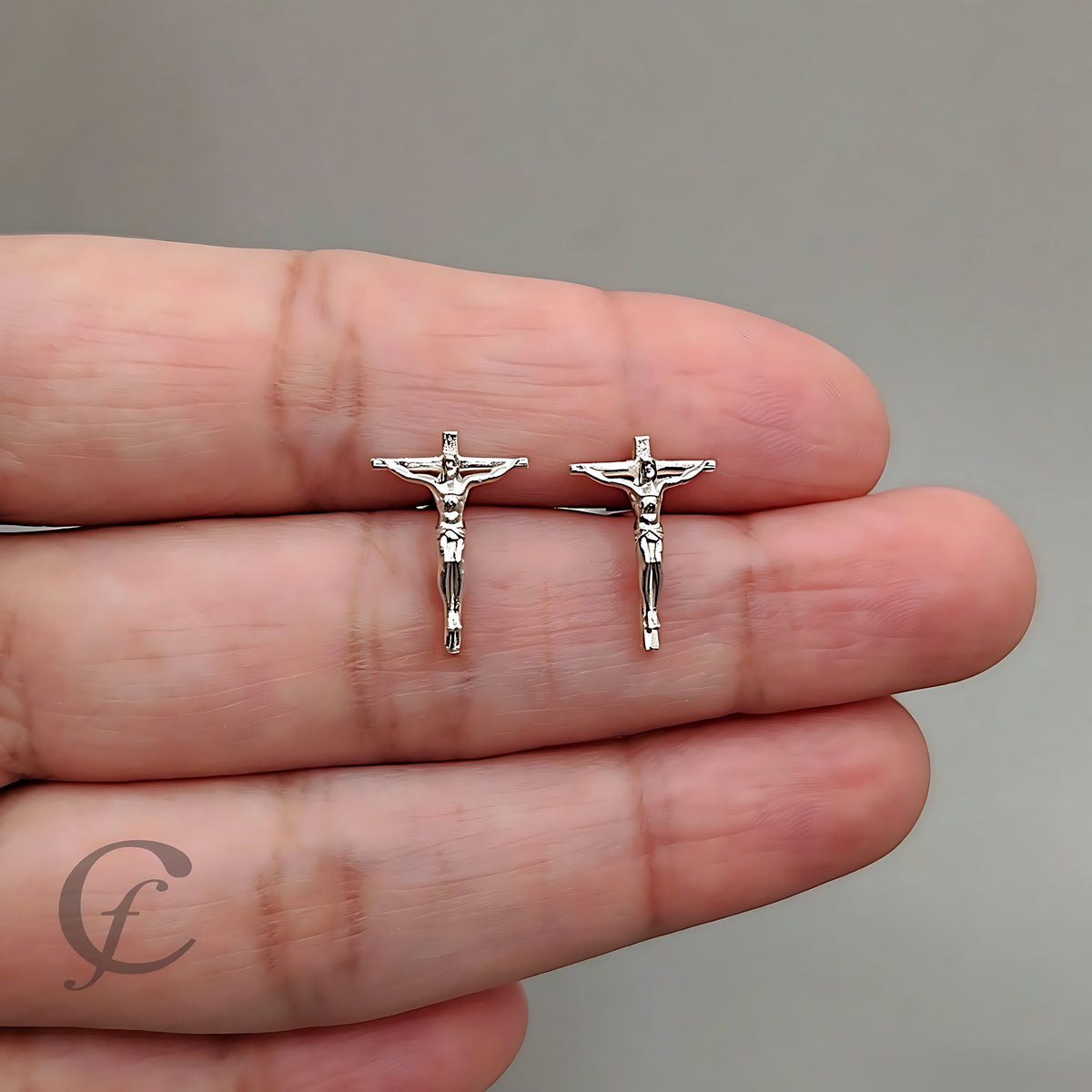 A pair of silver crucifix stud earrings displayed on a person's fingers against a neutral gray background.