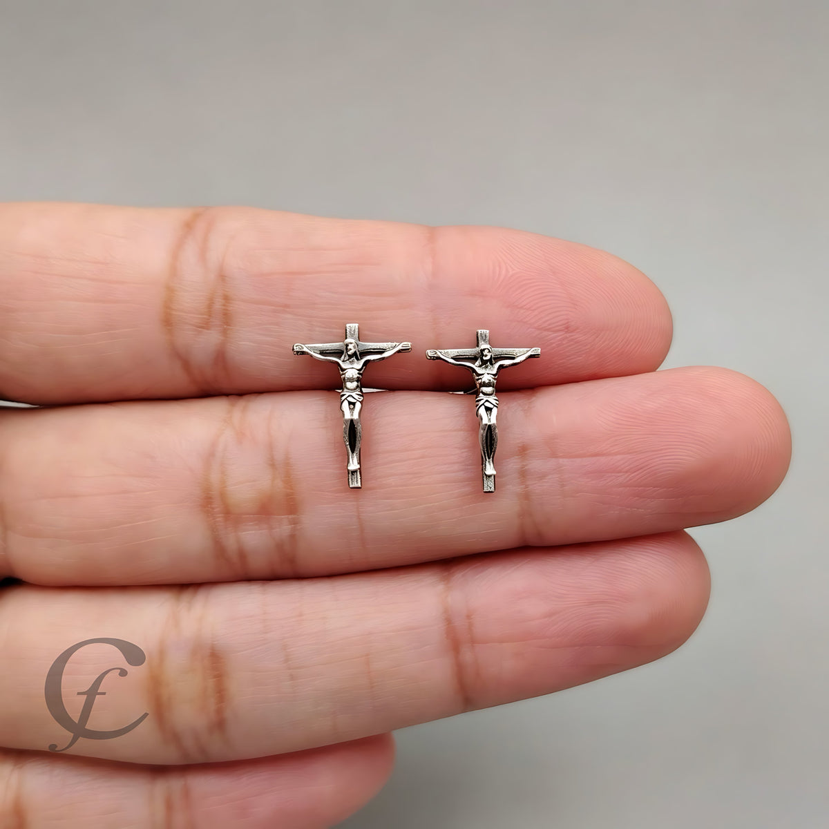 A pair of silver crucifix stud earrings held on a person's fingers against a plain light gray background.