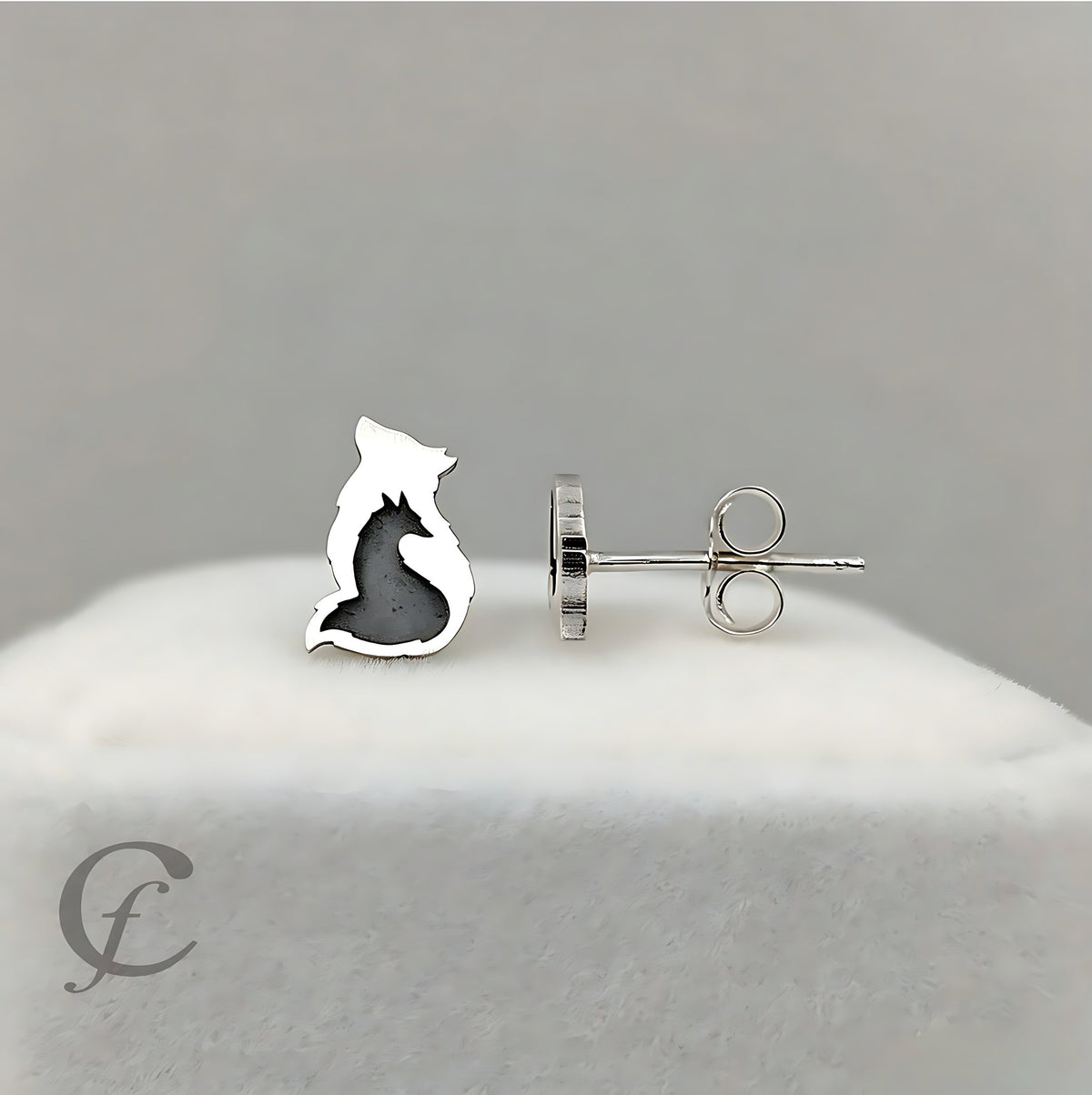 A pair of silver stud earrings shaped like a sitting cat, with one earring shown from the front and the other from the side/back. The cat silhouette has a dark gray inlay, and the earrings are displayed on a white textured stand against a neutral gray background.