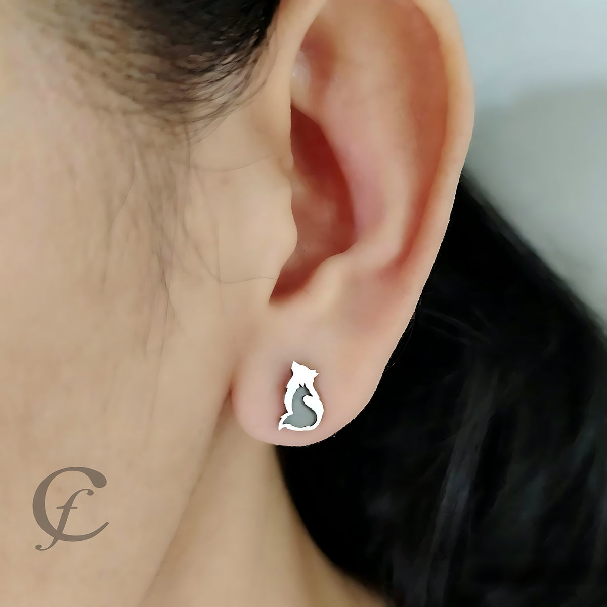 A close-up of a person's ear wearing a small silver stud earring shaped like a Wolf & Fox Silhouette with a gray fill.