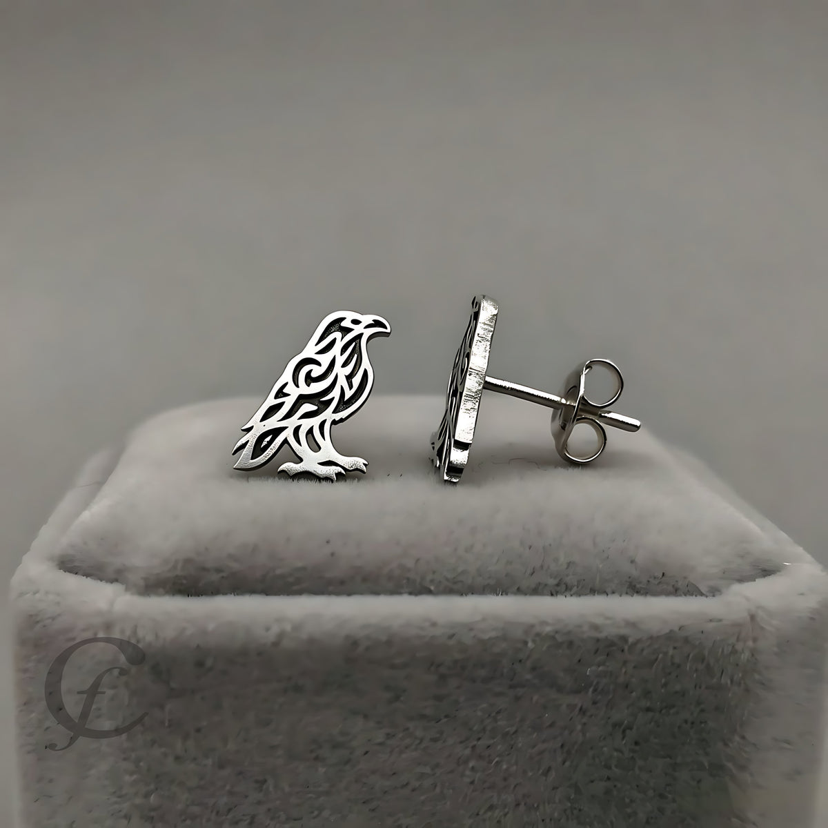 A pair of silver-toned raven stud earrings, one shown from the front and the other from the side/back, resting on a gray velvet jewelry box against a neutral gray background.