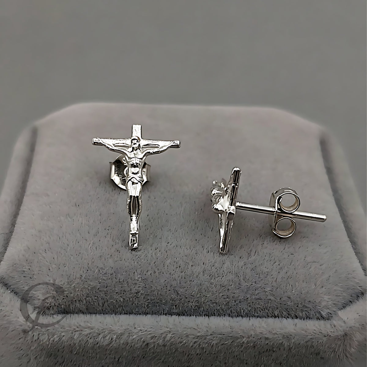 A pair of silver crucifix stud earrings displayed on a gray velvet jewelry box. One earring is shown from the front, depicting Jesus on the cross, while the other is shown from the side/back, displaying the post and butterfly backing.
