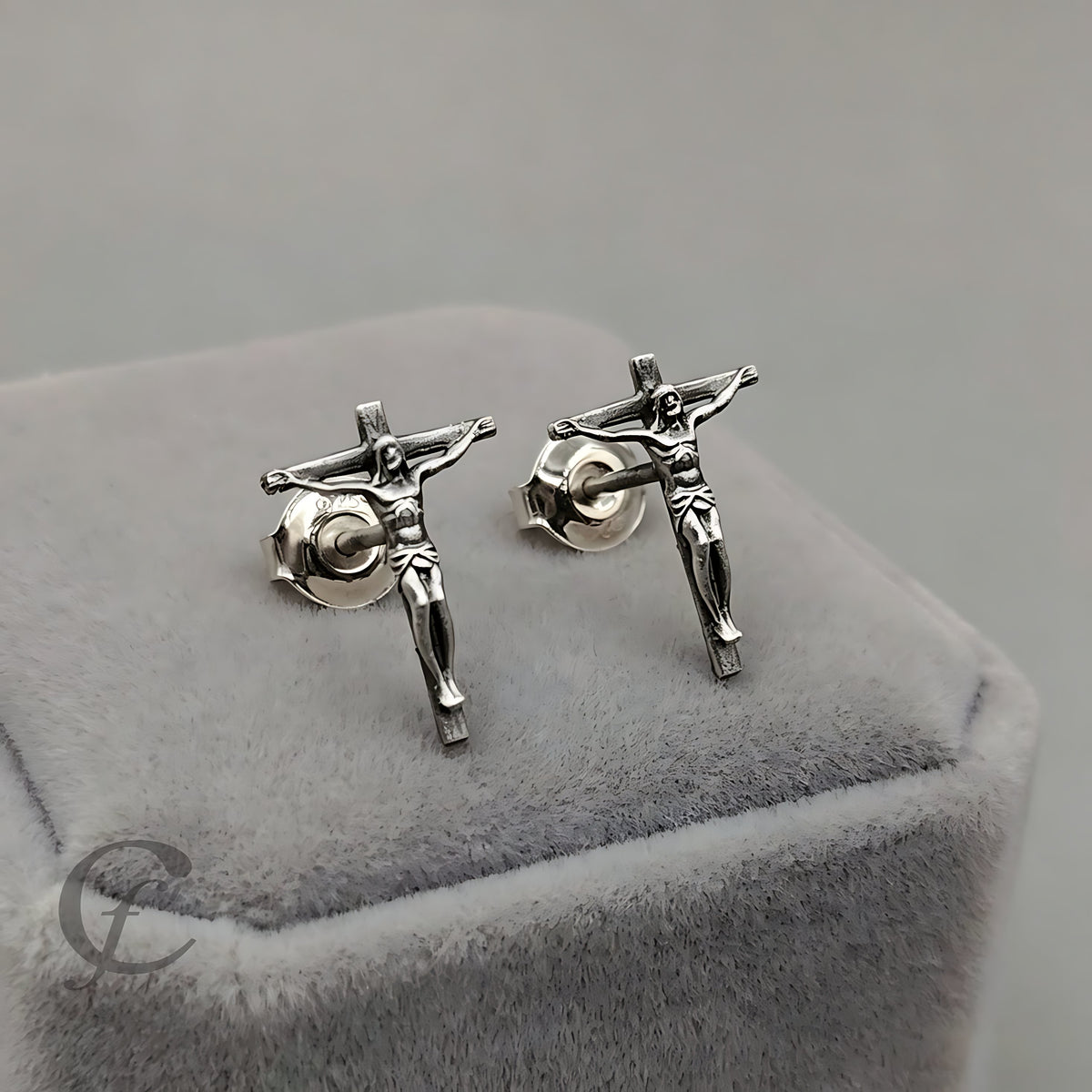 A pair of silver-toned crucifix stud earrings displayed on a gray velvet jewelry box against a neutral gray background.