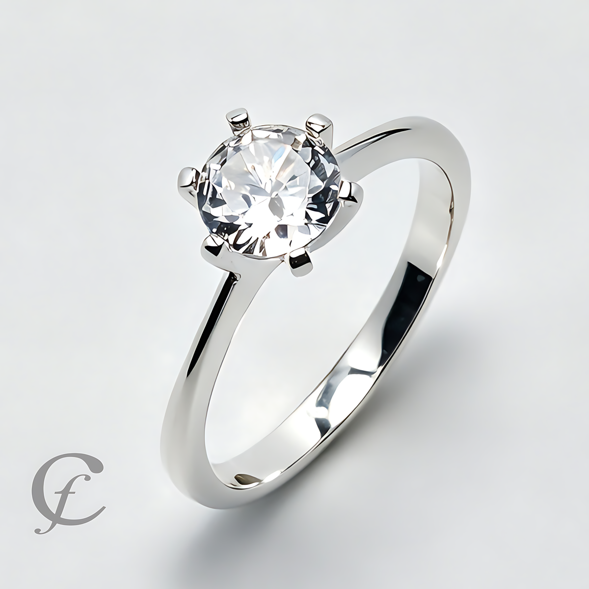 A silver solitaire engagement ring with a round brilliant-cut clear gemstone in a four-prong setting, shown on a light gray background with a watermark logo in the lower-left corner.
