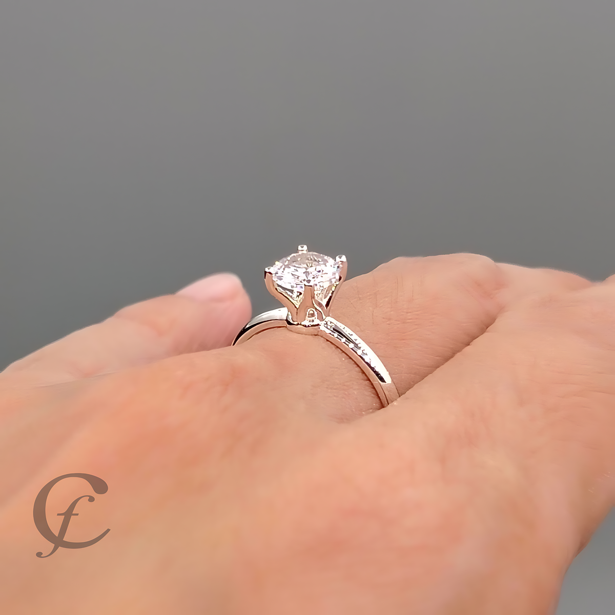 A close-up of a hand wearing a silver solitaire engagement ring with a cushion-cut clear gemstone. The ring is shown on a neutral gray background with a watermark logo in the bottom-left corner.