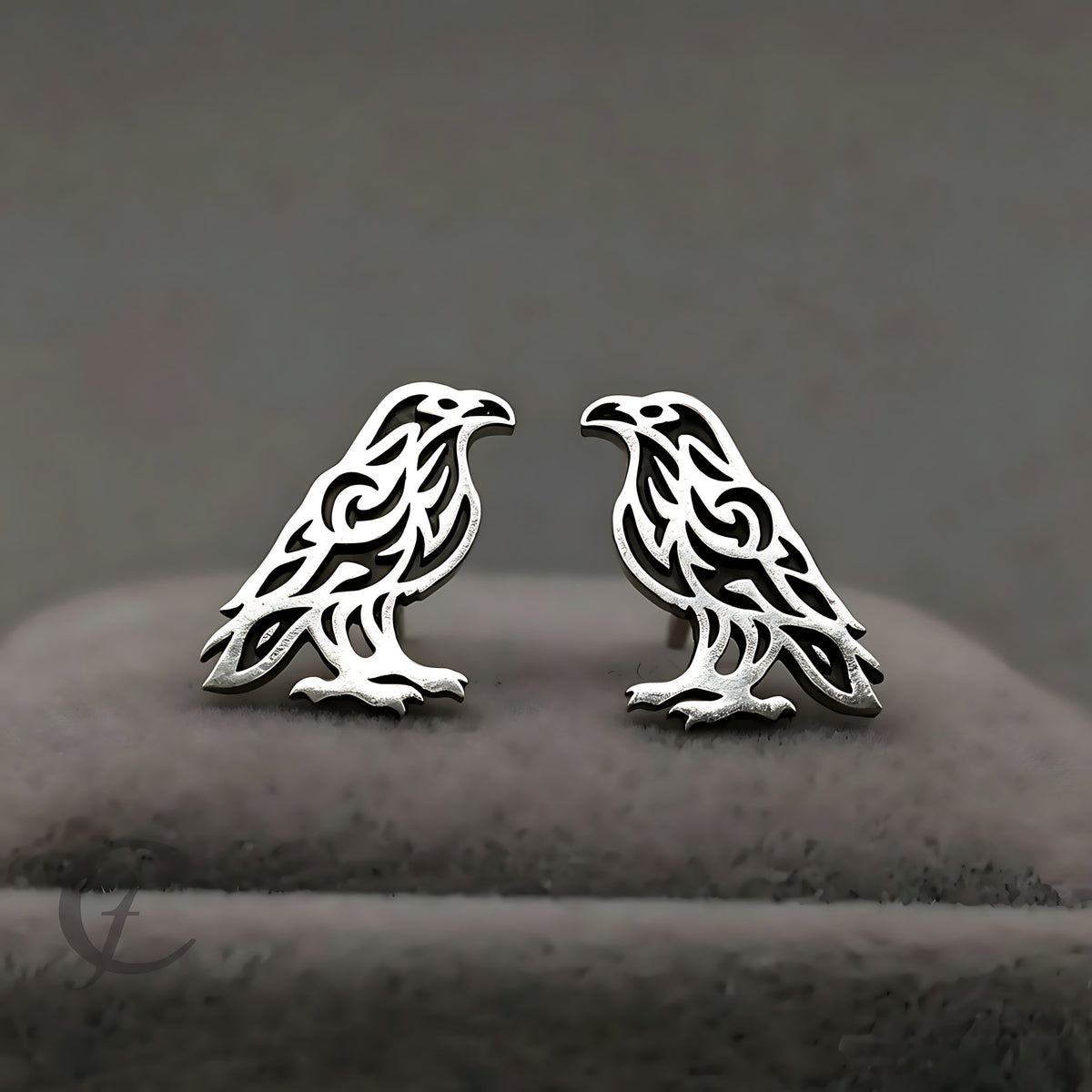A pair of silver-toned raven stud earrings with intricate cutout patterns, displayed on a dark gray velvet jewelry stand.