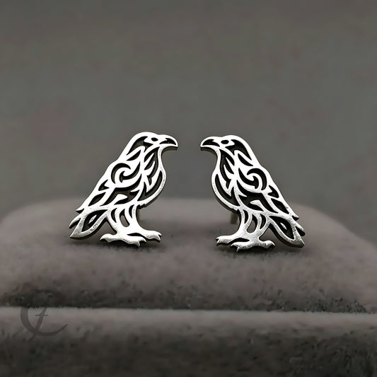 A pair of silver-toned raven stud earrings with intricate cutout patterns, displayed on a dark gray velvet jewelry stand.