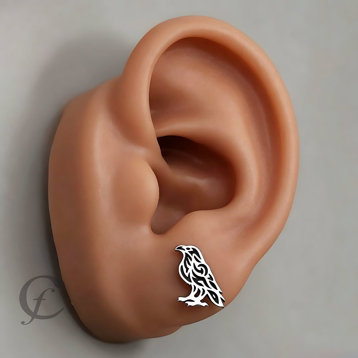 A silver-toned bird stud earring with a Celtic knot design, shown on the lobe of a realistic skin-toned ear against a neutral gray background.
