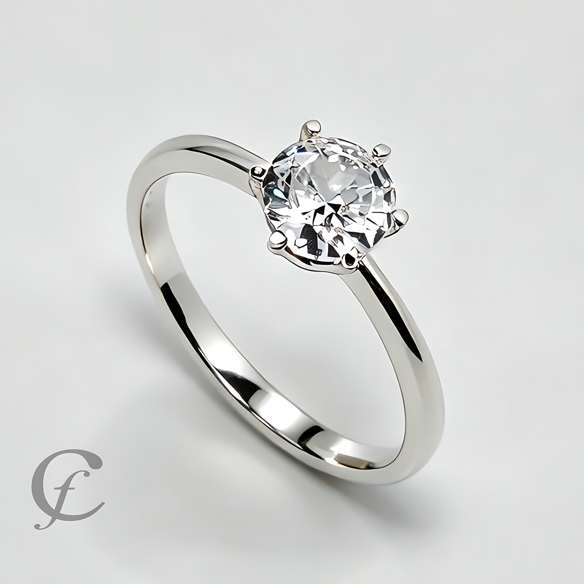 A classic solitaire engagement ring featuring a round brilliant-cut clear gemstone in a four-prong setting on a polished silver-colored band, shown on a light gray background with a watermark logo in the lower-left corner.