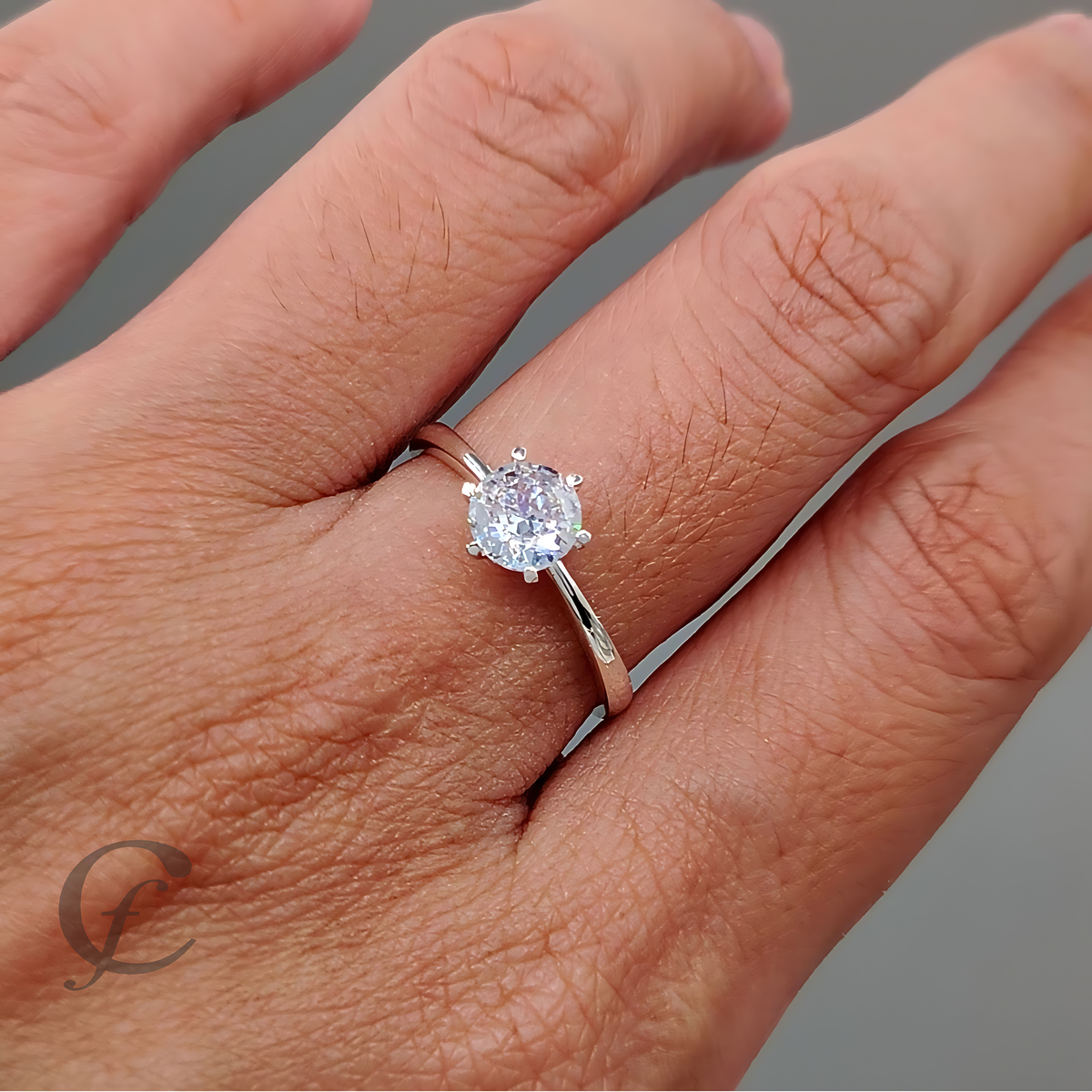 A classic solitaire engagement ring featuring a round brilliant-cut diamond set in a polished silver four-prong setting, on a matching silver band. The ring is photographed on a clean, light gray background with a soft shadow.