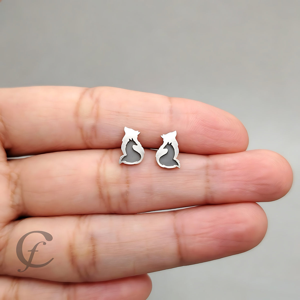 A pair of small silver stud earrings shaped like stylized Fox Wolf silhouettes with gray inlay, held between a person's fingers against a plain gray background.