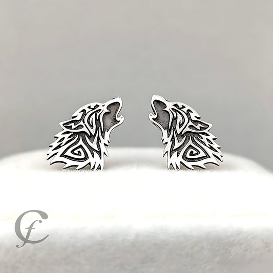 A pair of silver-toned wolf head stud earrings with intricate cutout detailing, displayed on a white textured surface against a soft gray background.