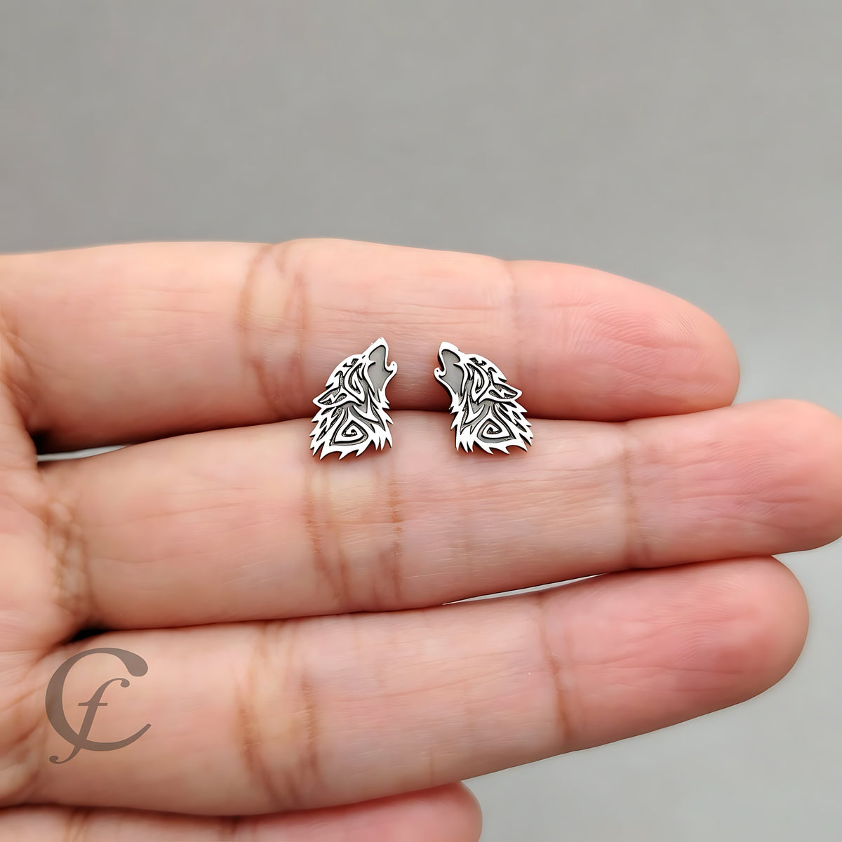 A pair of silver wolf-shaped stud earrings held on a person's fingers against a plain gray background.