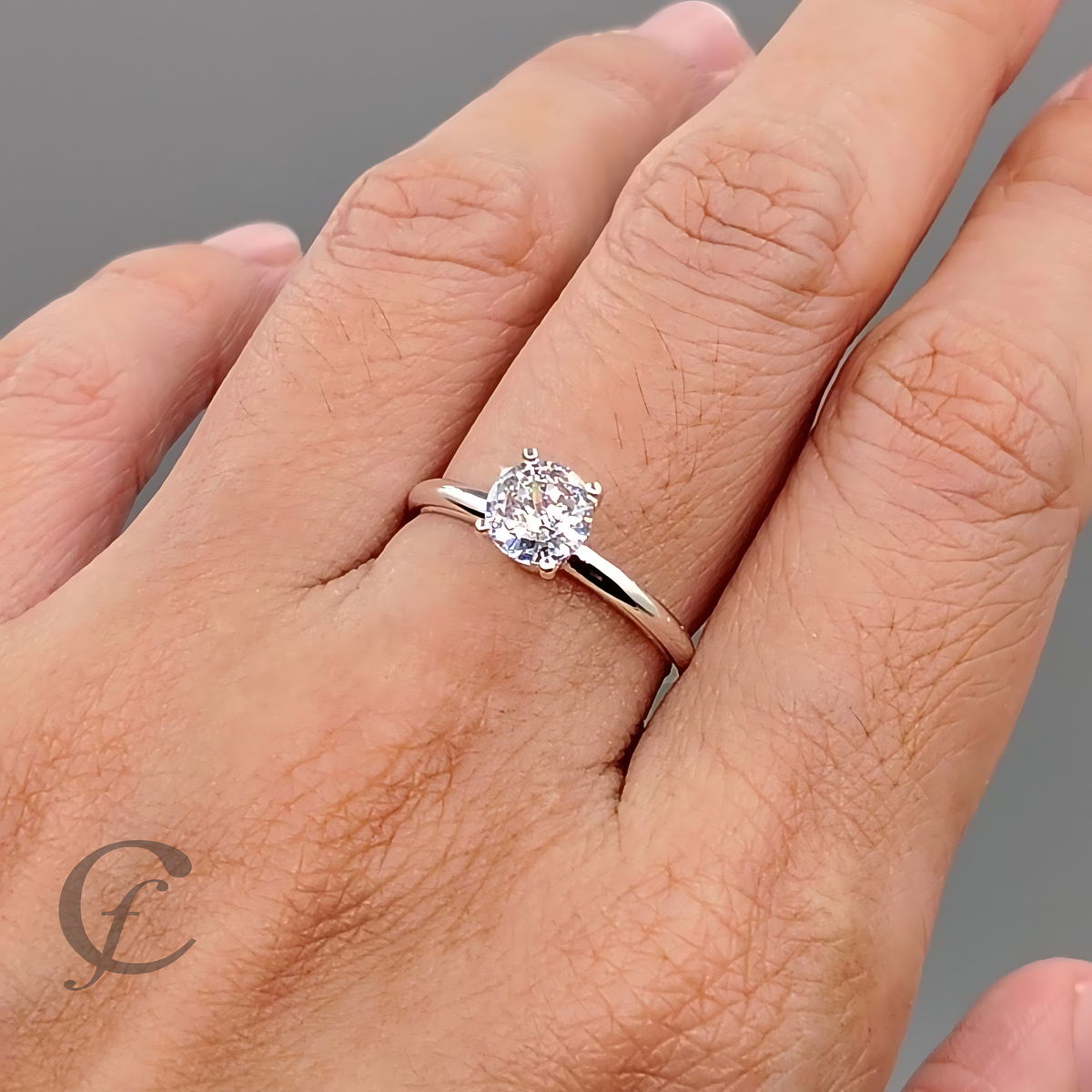 A silver solitaire engagement ring with a round-cut clear gemstone, shown worn on a person's ring finger against a neutral gray background.