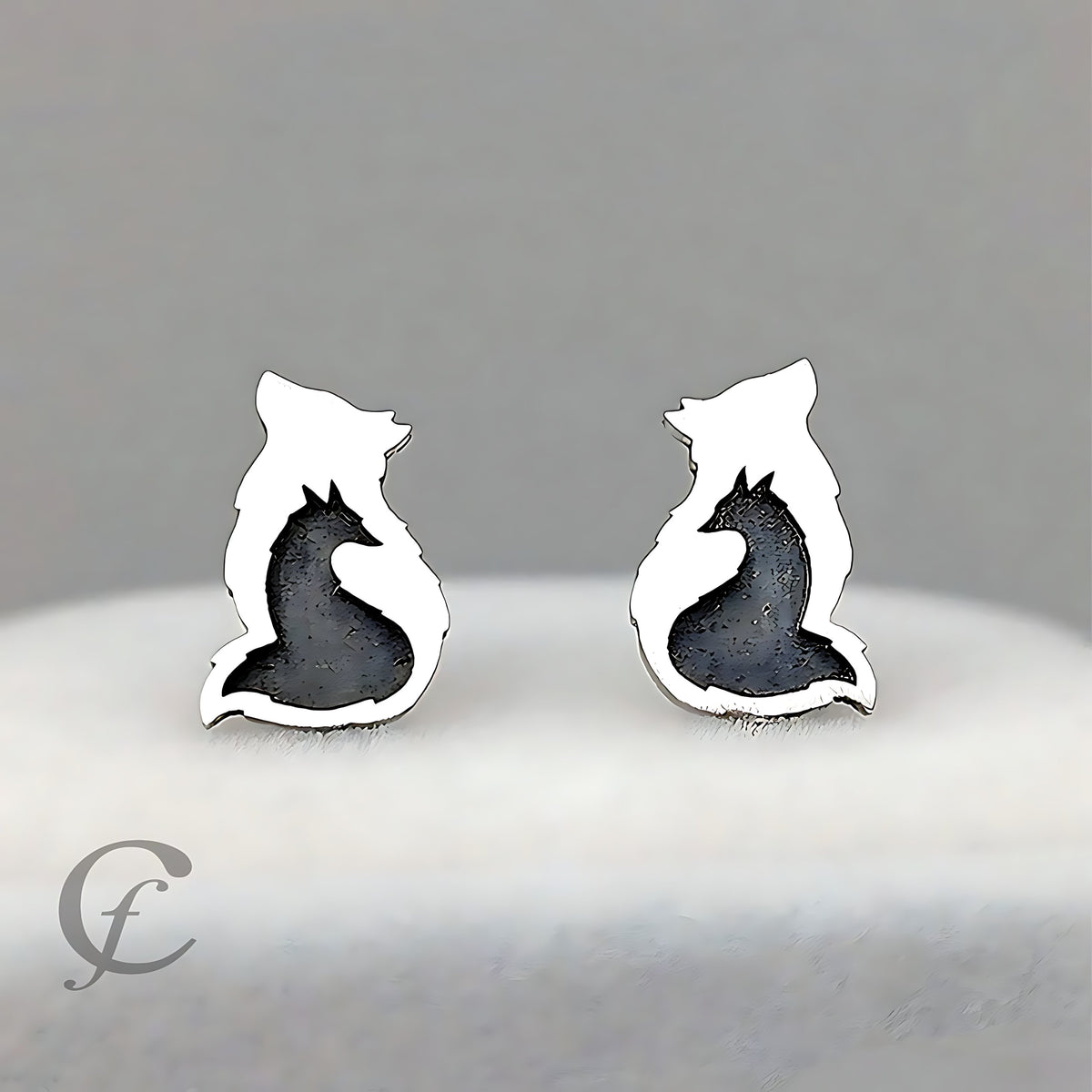 A pair of silver stud earrings shaped like foxes, featuring a polished silver outline with a textured dark gray/black inlay representing the fox's body, displayed on a soft white surface against a neutral gray background.