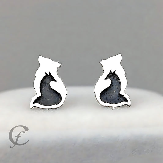 A pair of silver stud earrings shaped like foxes, featuring a polished silver outline with a textured dark gray/black inlay representing the fox's body, displayed on a soft white surface against a neutral gray background.