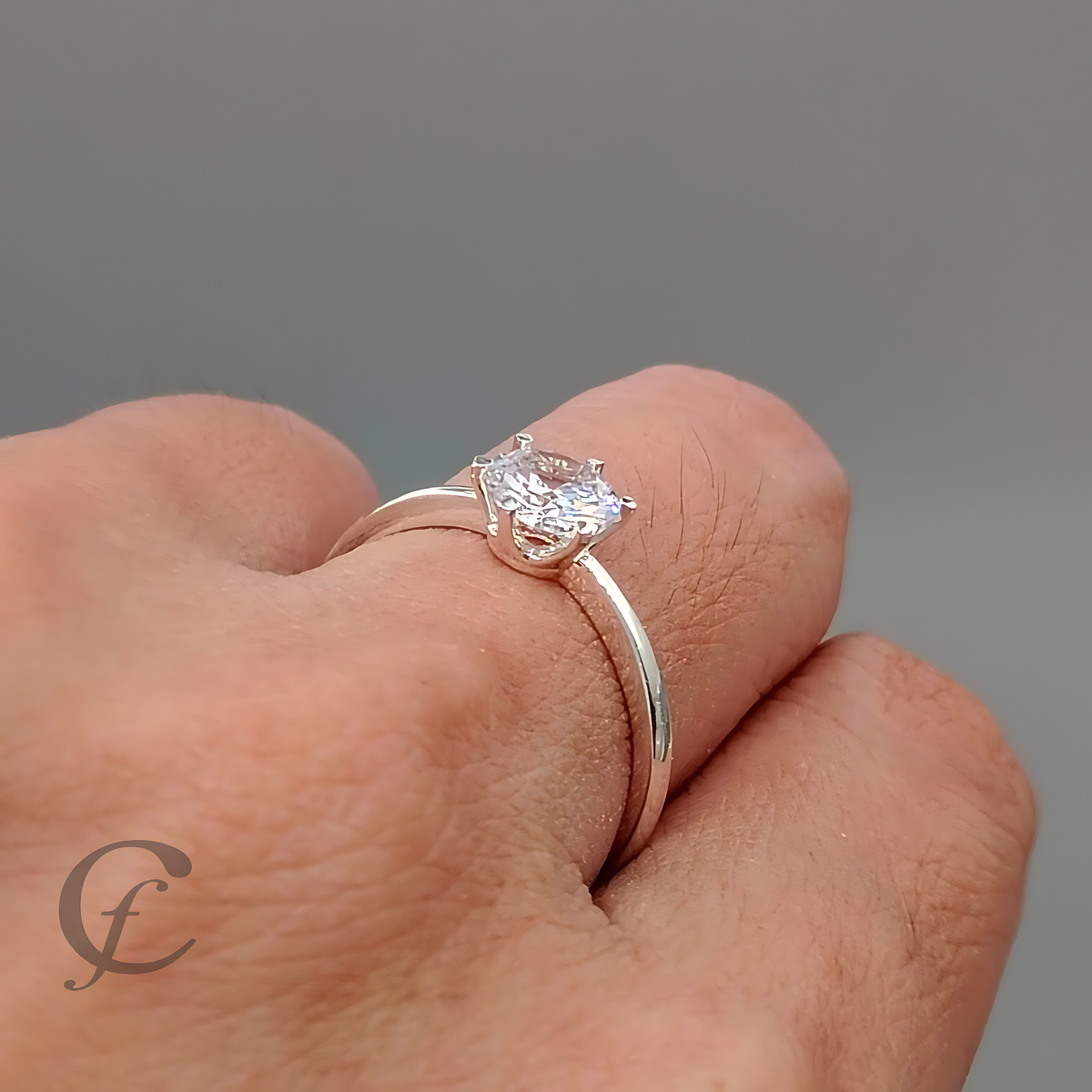 A silver solitaire ring with a clear heart-shaped gemstone worn on a person's finger, shown against a neutral gray background.