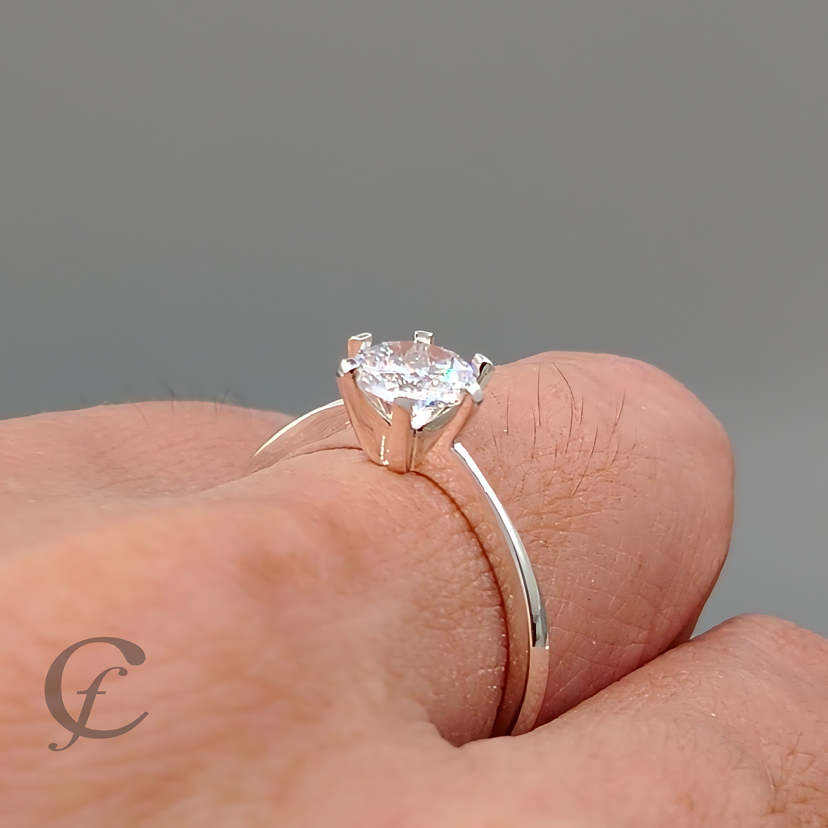 A silver solitaire engagement ring with a round brilliant-cut diamond set on a finger, shown against a neutral gray background.